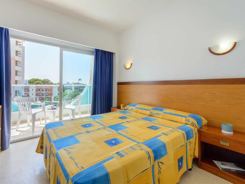 Lichte hotelkamer met tweepersoonsbed, houten hoofdeinde en balkon met uitzicht op nabijgelegen gebouwen.