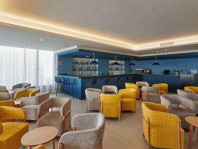 Salotto moderno in hotel con poltrone gialle e grigie, tavolini e scaffali blu.