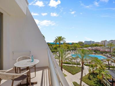Balcone d'hotel con tavolo e sedie, vista su piscina e palme sotto cielo azzurro.