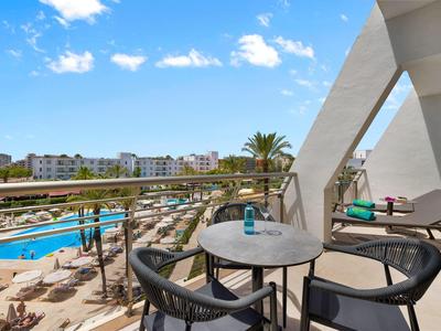 Balcone hotel con tavolo rotondo, sedie, vista piscina e edifici moderni sotto cielo azzurro.