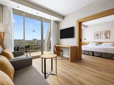 Camera d'hotel moderna con letto matrimoniale, divano, scrivania e balcone soleggiato.