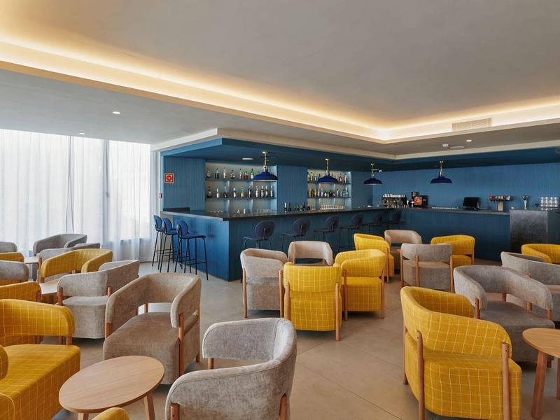 Salotto moderno in hotel con poltrone gialle e grigie, tavolini e scaffali blu.