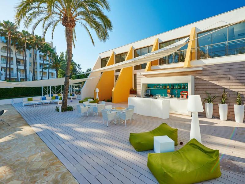 Terrazza moderna con sedute verdi e parete gialla accanto a un hotel soleggiato.