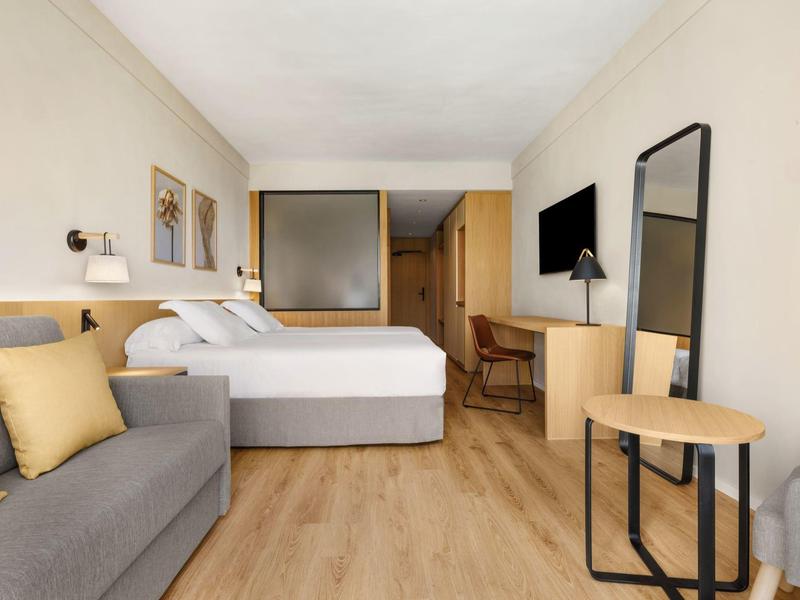 Camera d'hotel moderna con letto matrimoniale, divano grigio, TV e mobili in legno chiaro.