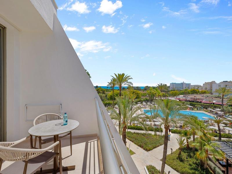 Balcone d'hotel con tavolo e sedie, vista su piscina e palme sotto cielo azzurro.