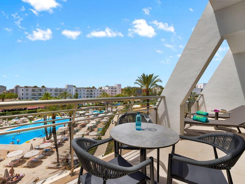 Balcone hotel con tavolo rotondo, sedie, vista piscina e edifici moderni sotto cielo azzurro.