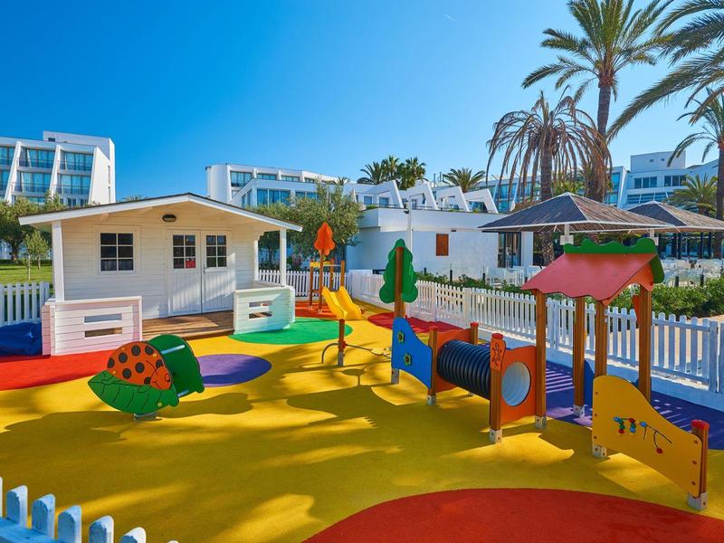 Area giochi colorata con superficie morbida, strutture per bambini e palme.