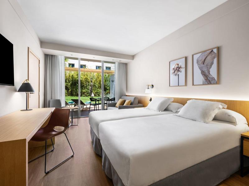 Camera d'hotel moderna con due letti, scrivania, sedia e balcone con vista sul verde.