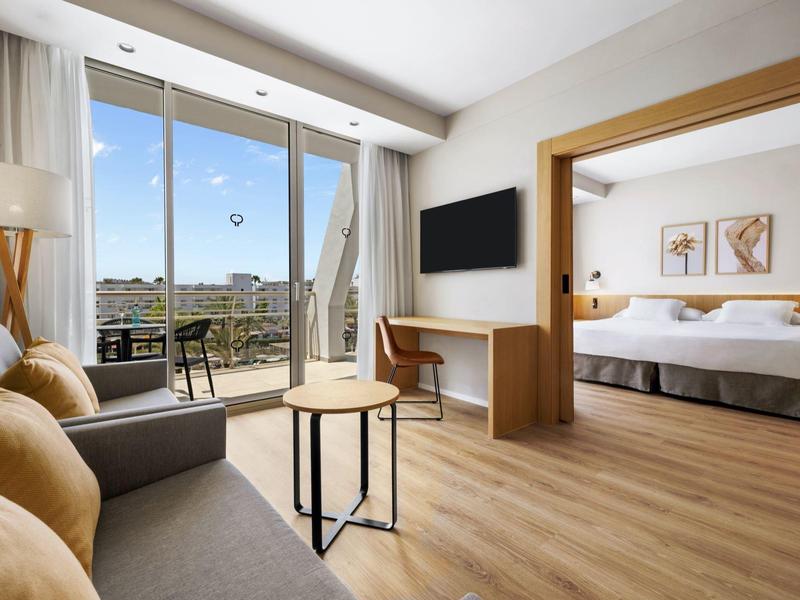 Camera d'hotel moderna con letto matrimoniale, divano, scrivania e balcone soleggiato.