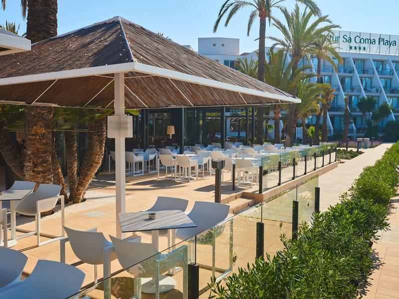 Terrazza esterna con tavoli, sedie e ombrelloni davanti a un edificio alberghiero e palme.