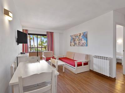 Světlý hotelový pokoj s jídelním koutem, dvojlůžkem, televizí na stěně, dřevěnou podlahou a výhledem na palmy.