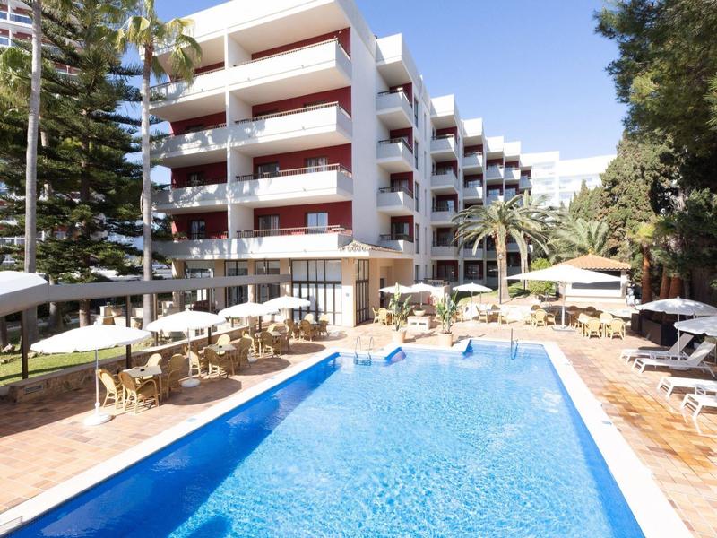 Moderní venkovní bazén s lehátky a slunečníky vedle vícepodlažní hotelové budovy.