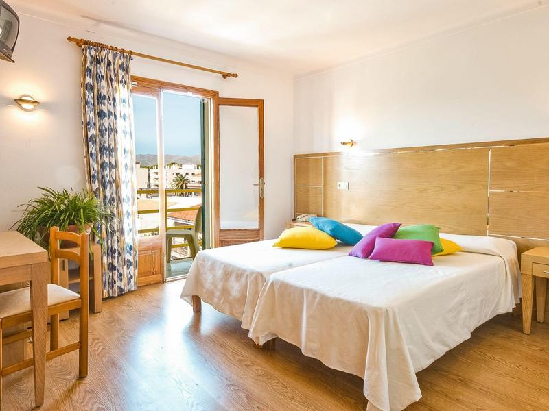 Lichte hotelkamer met tweepersoonsbed, kleurrijke kussens en balkon met uitzicht buiten.