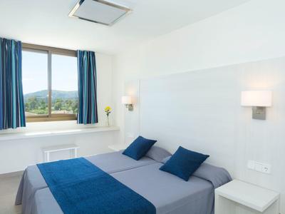 Jasny pokój hotelowy z podwójnym łóżkiem, niebieskimi zasłonami i widokiem na przyrodę.