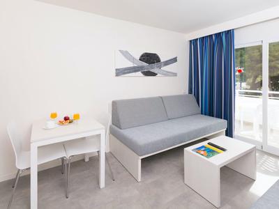 Nowoczesny pokój hotelowy z szarą sofą, białym stołem, krzesłami i otwartym balkonem.