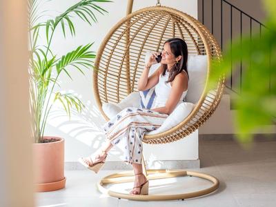 Kobieta na wiszącym krześle z rattanu, pije coś w pomieszczeniu obok zielonej rośliny.