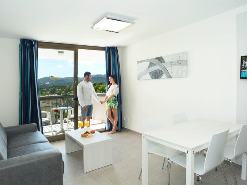 Jasne mieszkanie z szarą sofą, białym stołem i balkonem z widokiem, para patrzy na zewnątrz.