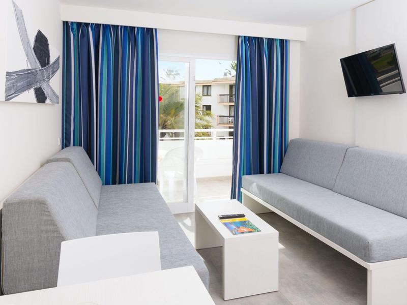 Hotelowy salon z dwoma szarymi sofami, stolikiem i telewizorem na ścianie.