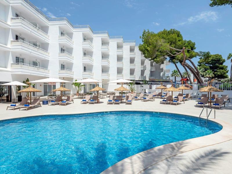 Niebieski basen z leżakami i parasolami przed białym hotelem pod jasnym niebem.