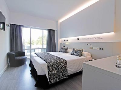 Bright hotel room with double bed, gray armchair, and balcony with curtains.