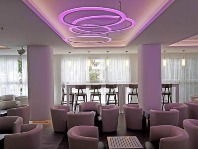 Modern hotel lounge with pink lighting, armchairs, and high tables next to large windows.