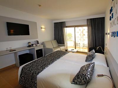 Modern hotel room with two beds, desk, television, and balcony with outdoor view.