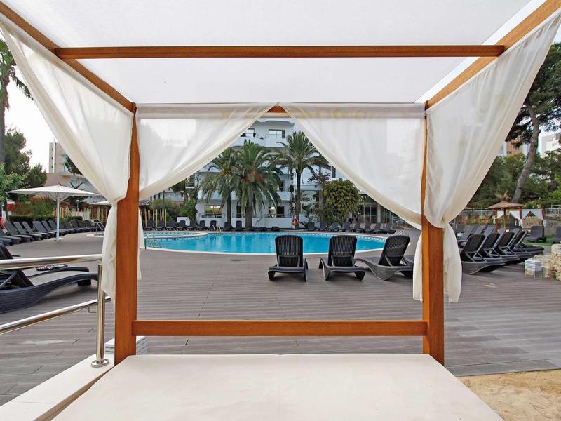 View from a covered pool lounger overlooking a pool area with sunbeds and palm trees.