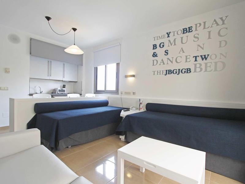 Modern hotel room with two single beds, sofa, and kitchenette.