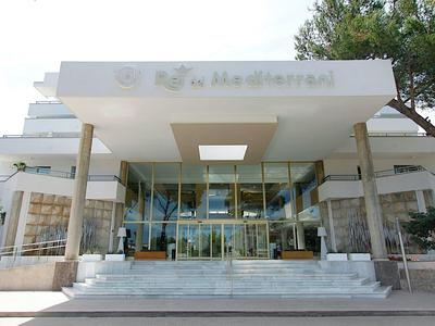 Eingangsbereich eines modernen Gebäudes mit Glasfront, weißen Säulen und Bäumen im Hintergrund.