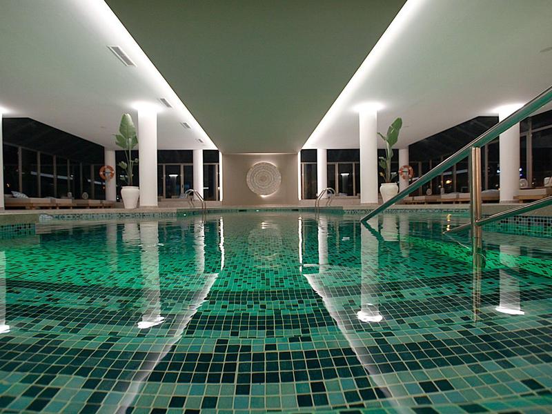 Innenpool mit grün-blau gemusterten Fliesen, weißen Säulen und indirekter Deckenbeleuchtung.