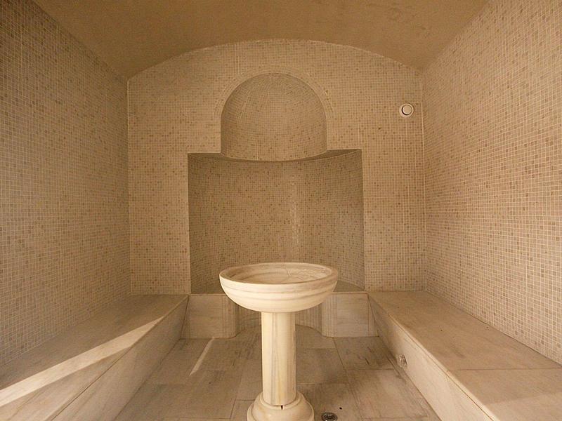 Innenraum einer Hammam-Sauna mit Mosaikwänden, Steinbänken und zentralem Wasserbecken.