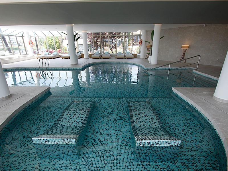 Innen gep?egtes Schwimmbad mit raumhohen Fenstern und Wassersitzbänken aus Mosaikfliesen.