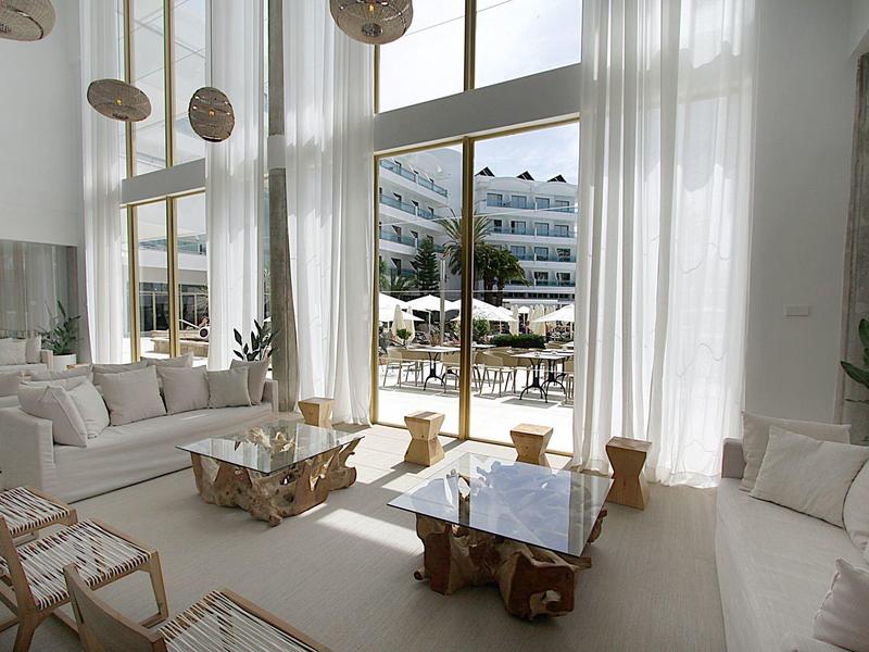 Helles Wohnzimmer mit hohen Fenstern, weißen Vorhängen, Holz- und Glasscouchtischen, bequemen Sesseln.