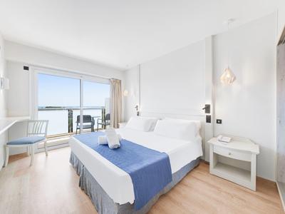 Helles Hotelzimmer mit Doppelbett, blauer Tagesdecke und Balkon mit Meerblick.