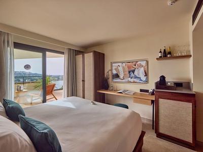 Schlafzimmer mit Doppelbett, Balkon, Stadtblick, Fernseher an der Wand und minimaler Dekoration.