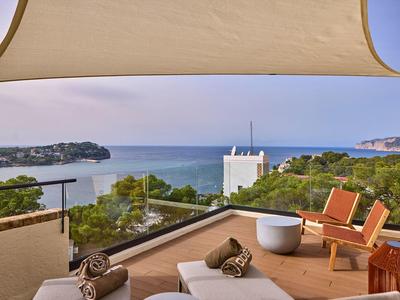 Terrasse mit Liegen, Stühlen, Blick auf Meer und grüne Hügel, warmer Holz- und Sandfarbton.