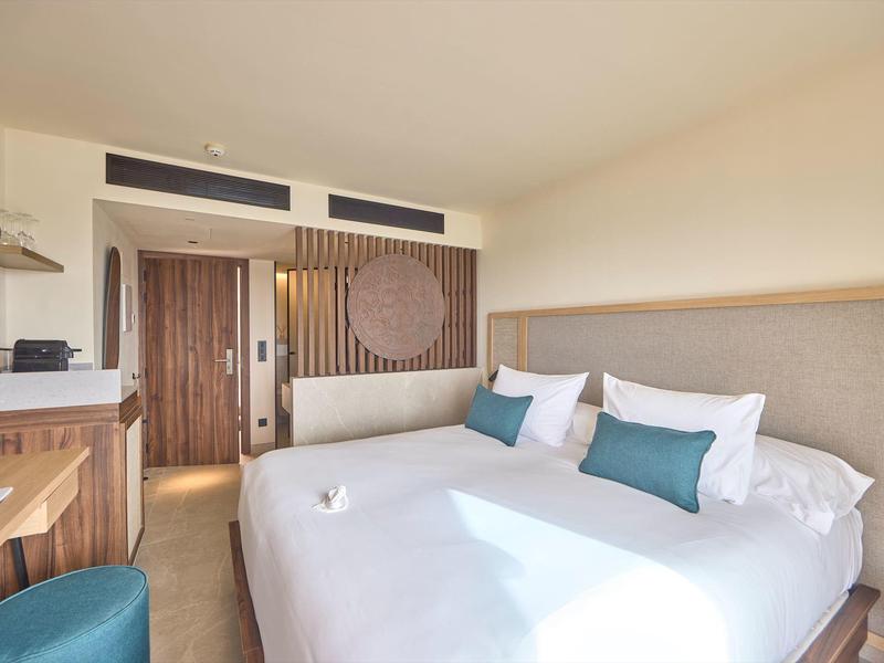 Modernes Hotelzimmer mit großem Bett, hellen Wänden, Holzmöbeln und blau-grünen Akzenten.