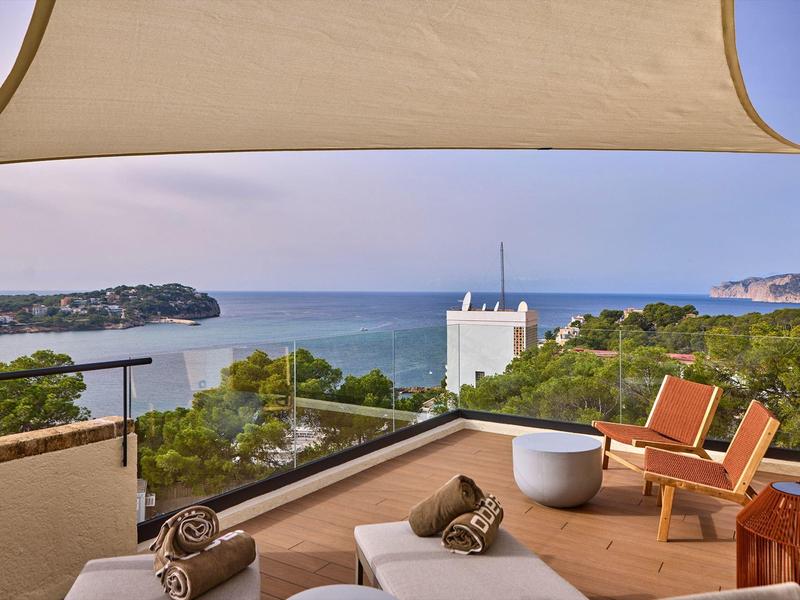 Terrasse mit Liegen, Stühlen, Blick auf Meer und grüne Hügel, warmer Holz- und Sandfarbton.