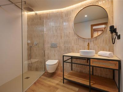 Baño moderno con espejo redondo, inodoro de pared y ducha abierta con suelo de madera.