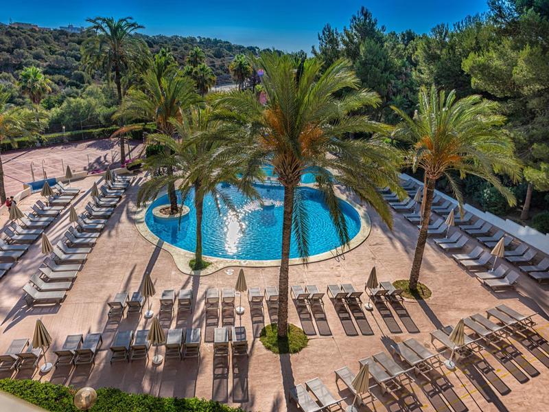 Piscina redonda con palmeras y muchas tumbonas en un complejo hotelero soleado.