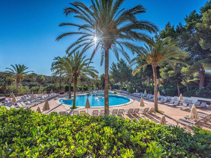 Piscina soleada de hotel con palmeras, tumbonas y cielo azul claro al fondo.