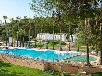 Buitenzwembad omringd door ligstoelen, palmbomen en witte cabana's in een zonnige tuin.