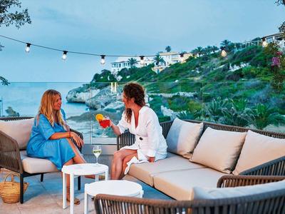 Twee vrouwen genieten van drankjes op een terras met comfortabele zitplaatsen en uitzicht op de kust bij schemer.