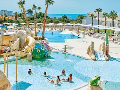 Buitenzwembad van hotel met glijbanen, palmbomen en uitzicht op zee, kinderen zwemmen en spelen.