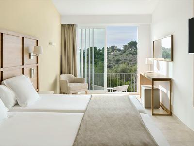 Lichte hotelkamer met twee bedden, fauteuil, bureau en balkon met uitzicht op groen.