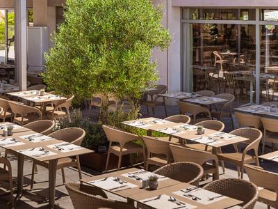 Outdoor restaurant terrace with tables, chairs, and a green tree plant, sunny.