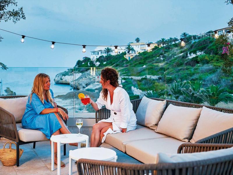 Twee vrouwen genieten van drankjes op een terras met comfortabele zitplaatsen en uitzicht op de kust bij schemer.