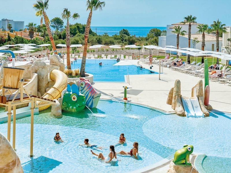 Buitenzwembad van hotel met glijbanen, palmbomen en uitzicht op zee, kinderen zwemmen en spelen.