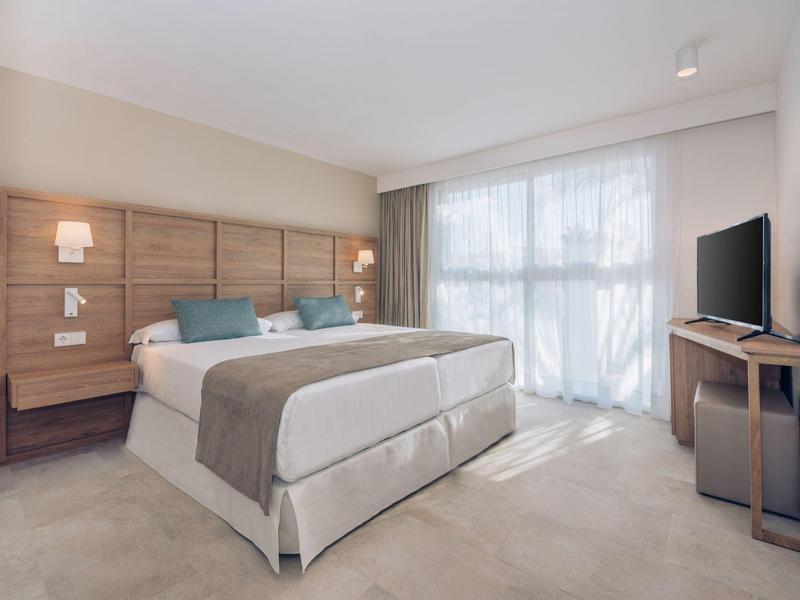 Lichte hotelkamer met groot bed, houten hoofdeinde, tv en vitrage die natuurlijk licht binnenlaat.