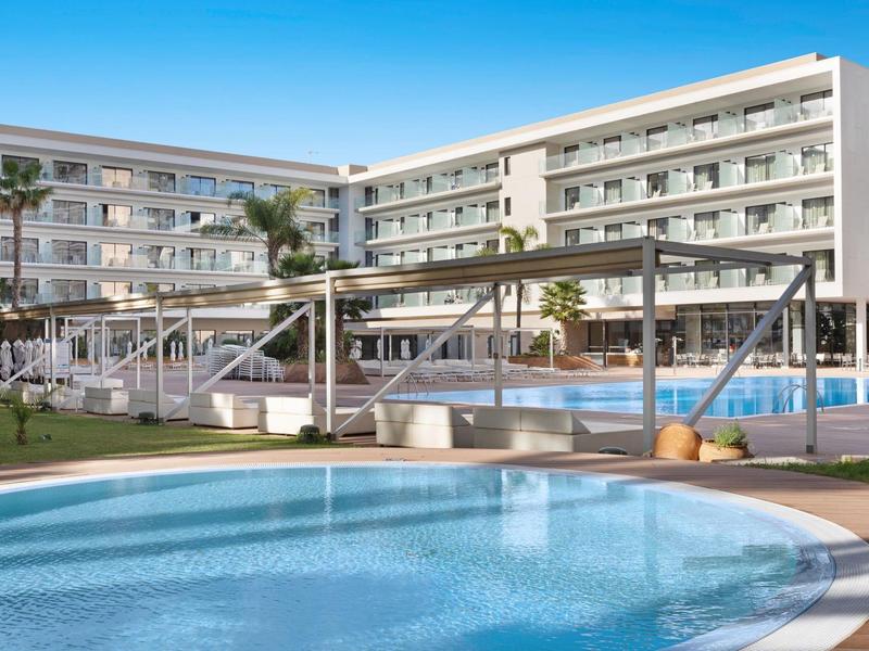 Complejo hotelero moderno con piscina, áreas para tomar el sol y puente de madera bajo el cielo despejado.